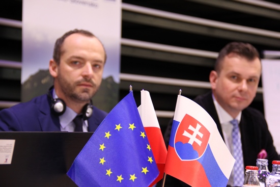 Na pierwszym planie trzy flagi: polska, słowacka i unijna. Za nimi dwie osoby siedzące przy stole.